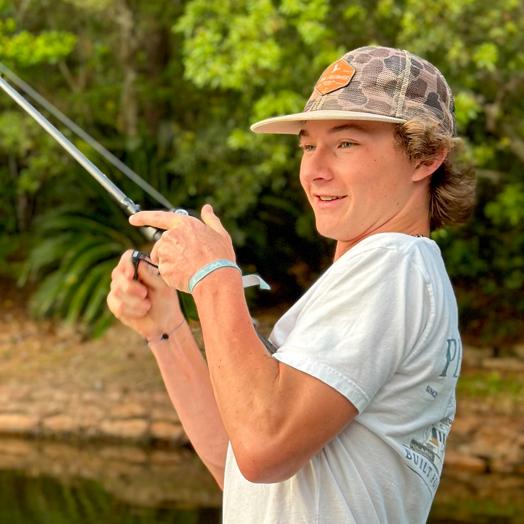 A young man wearing a Fowl Follower Mesh Camo Hat, holding a fishing rod and a fishing pole.